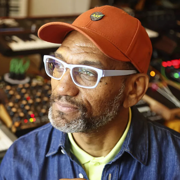 King Britt wearing an orange baseball cap with white frame glasses and a neon yellow/green shirt with a denim button up jacket while looking off to the left side in his studio with keyboards and synthesizers in the background. 