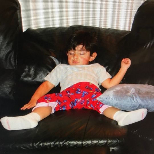 A picture of a boy (maybe around 8-years-old) who is asleep on the couch. He has short black hair and wears a white t-shirt and red Spider-man pajama pants and socks. 